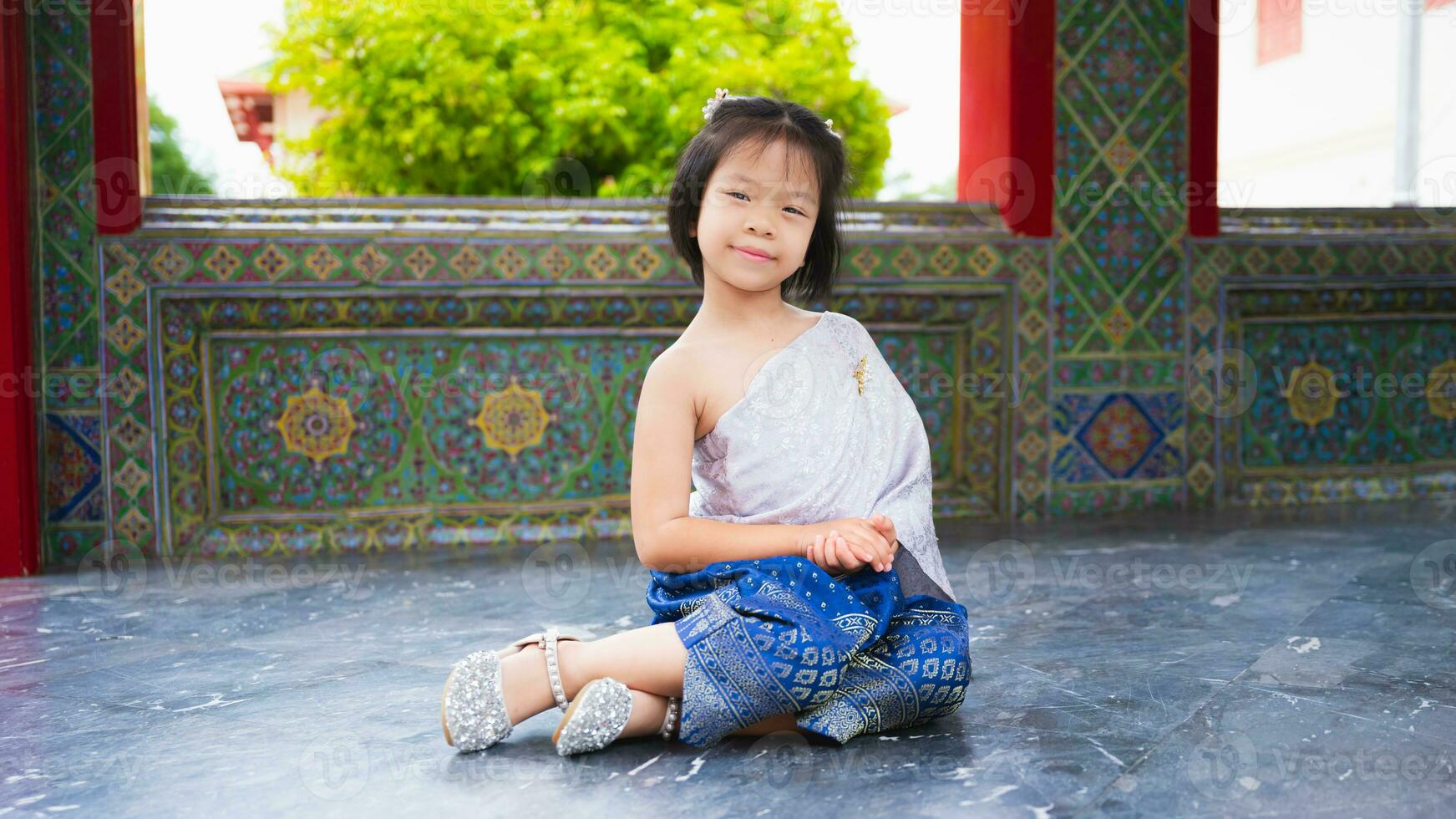 schattig meisje draagt Thais jurk met zilver sjerp en blauw lendendoek bijgesneden met goud. Aziatisch kind bezoeken tempel in Thailand naar zien kunst en cultuur met haar familie. kind squats zitten Aan de verdieping en glimlacht. foto