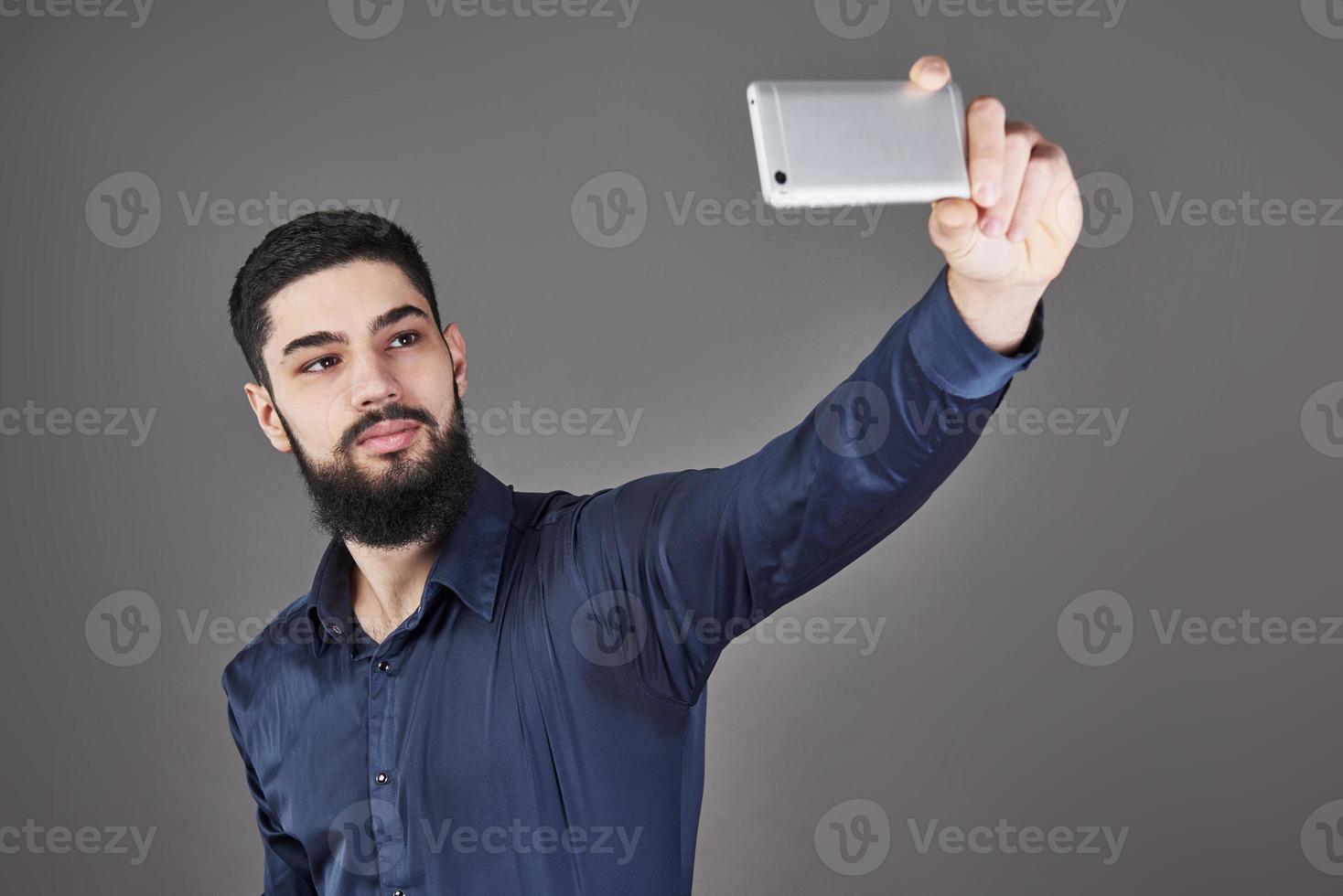 jonge, bebaarde hipster zakenman praten selfie foto met slimme telefoon glimlachen en kijken naar telefoon tegen grijze studio achtergrond