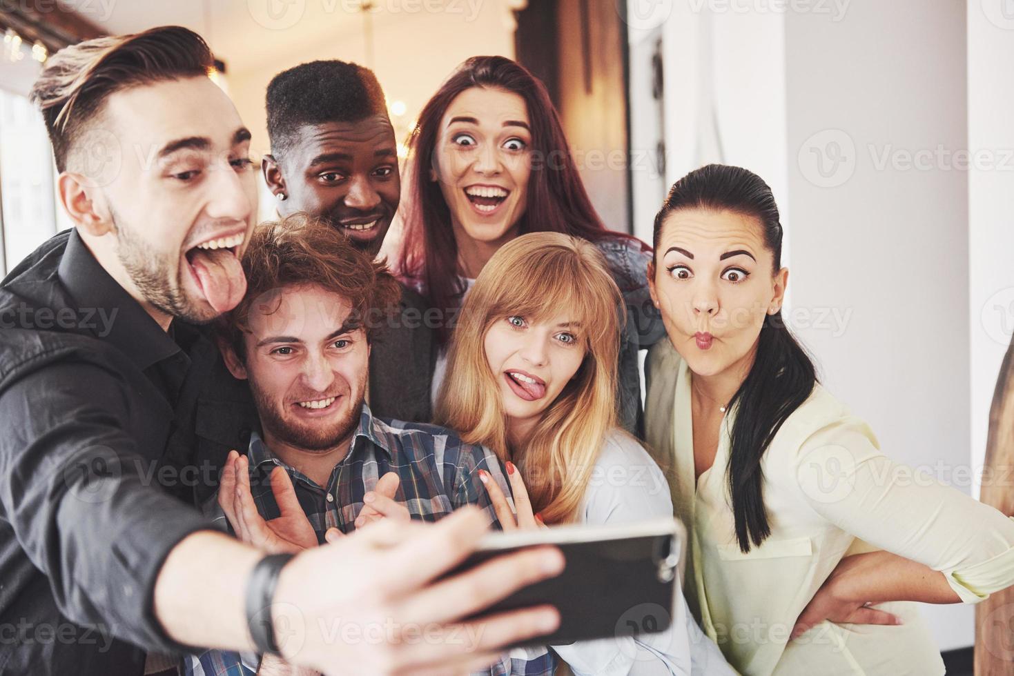 vrienden die plezier hebben in restaurant. twee jongens en vier meisjes drinken selfie maken, vredesteken maken en lachen. op de voorgrond vrouw met slimme telefoon. dragen allemaal vrijetijdskleding foto