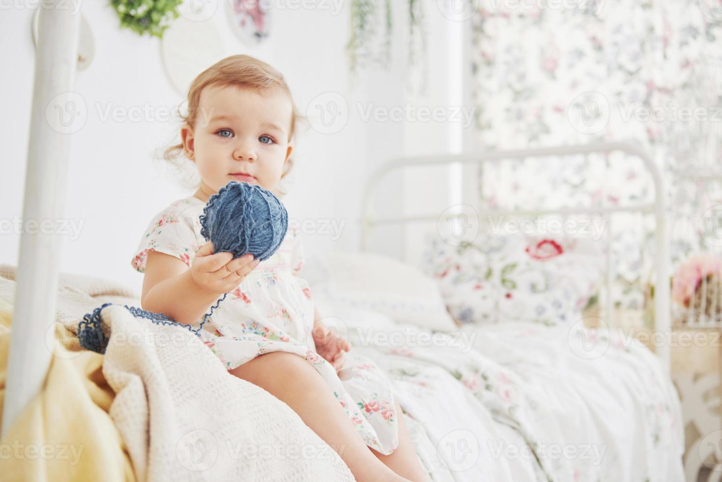 kindertijd concept. meisje van de baby in schattige jurk aanbrengen op bed spelen met speelgoed door het huis. witte vintage kinderkamer foto