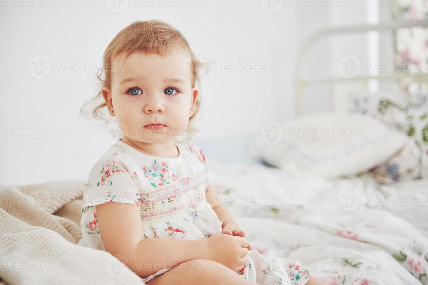 kindertijd concept. meisje van de baby in schattige jurk aanbrengen op bed spelen met speelgoed door het huis. witte vintage kinderkamer foto