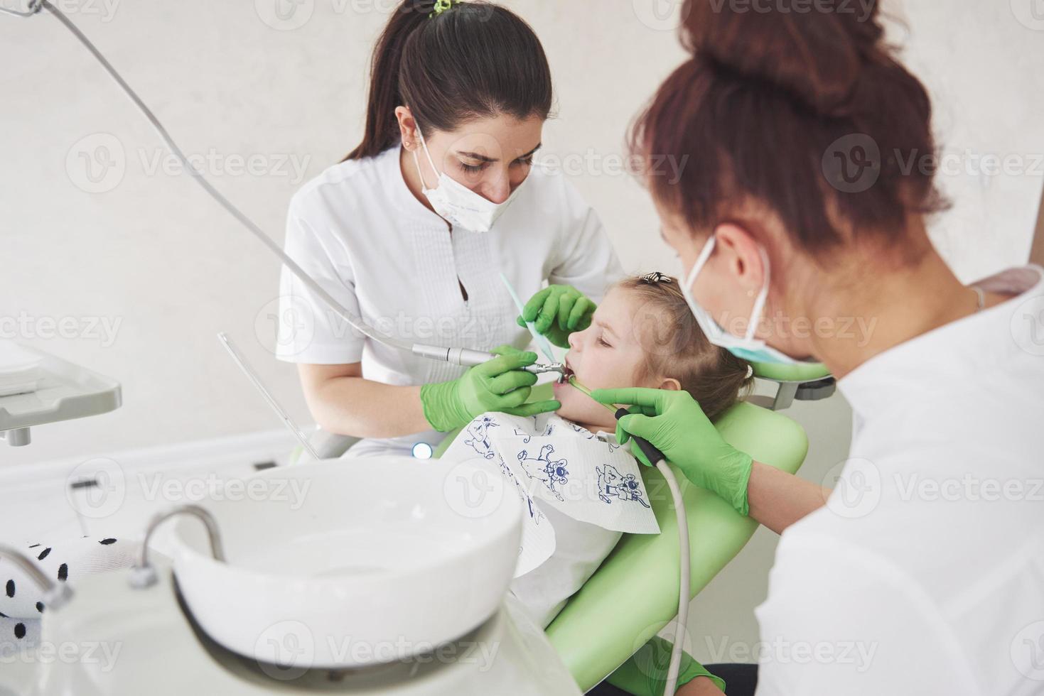 handen van onherkenbare pediatrische tandarts en assistent die onderzoeksprocedure maken voor lachend schattig klein meisje zittend op een stoel in het ziekenhuis foto