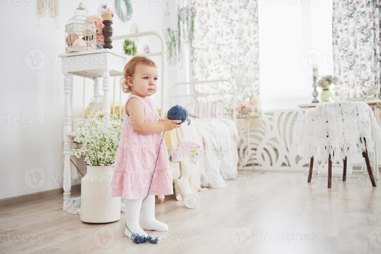 kindertijd concept. babymeisje in schattig jurkje spelen met gekleurde draad. witte vintage kinderkamer foto