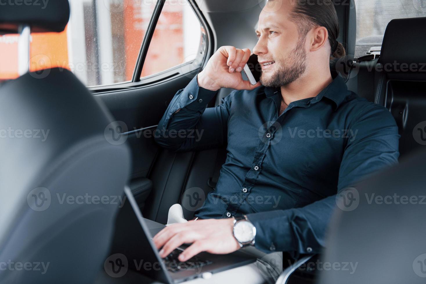 een jonge zakenman die aan een laptop werkt en aan de telefoon praat terwijl hij in de auto zit. werkt in beweging, waardeert zijn tijd foto