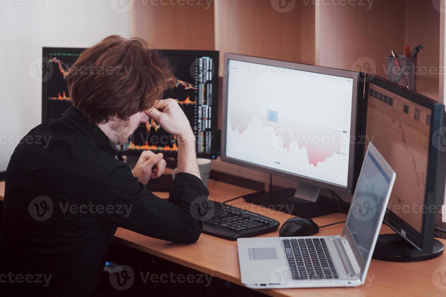 stressvolle dag op kantoor. jonge zakenman hand in hand op zijn gezicht zittend aan de balie in creatief kantoor. beurshandel forex financiën grafisch concept foto