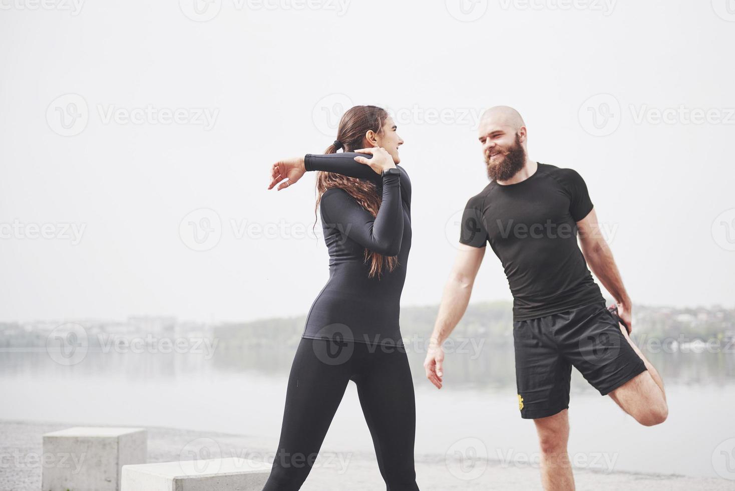 fitness paar die zich uitstrekt buiten in het park in de buurt van het water. jonge, bebaarde man en vrouw die 's ochtends samen trainen foto