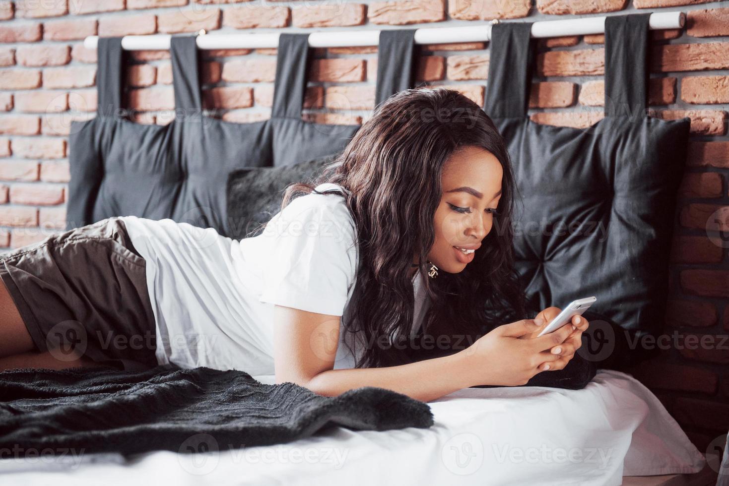 portret van mooie vrouw wakker in haar bed en kijkt in de telefoon. controleer sociale netwerken, stuur sms. het meisje draagt een t-shirt foto