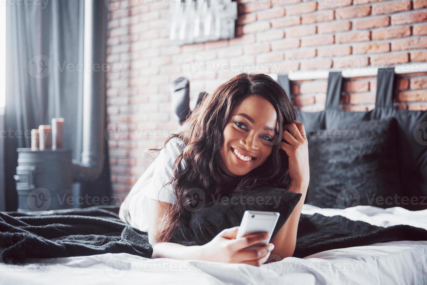 portret van mooie vrouw wakker in haar bed en kijkt in de telefoon. controleer sociale netwerken, stuur sms. het meisje draagt een t-shirt foto