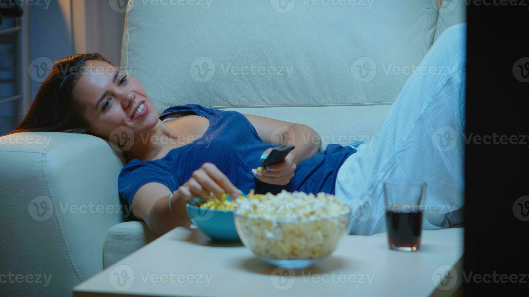 jong vrouw in pyjama's resting aan het liegen Aan bankstel in voorkant TV lachend. Vrolijk, geamuseerd, eenzaam dame genieten van de avond zittend Aan comfortabel sofa aan het kijken televisie aan het eten popcorn en snacks foto