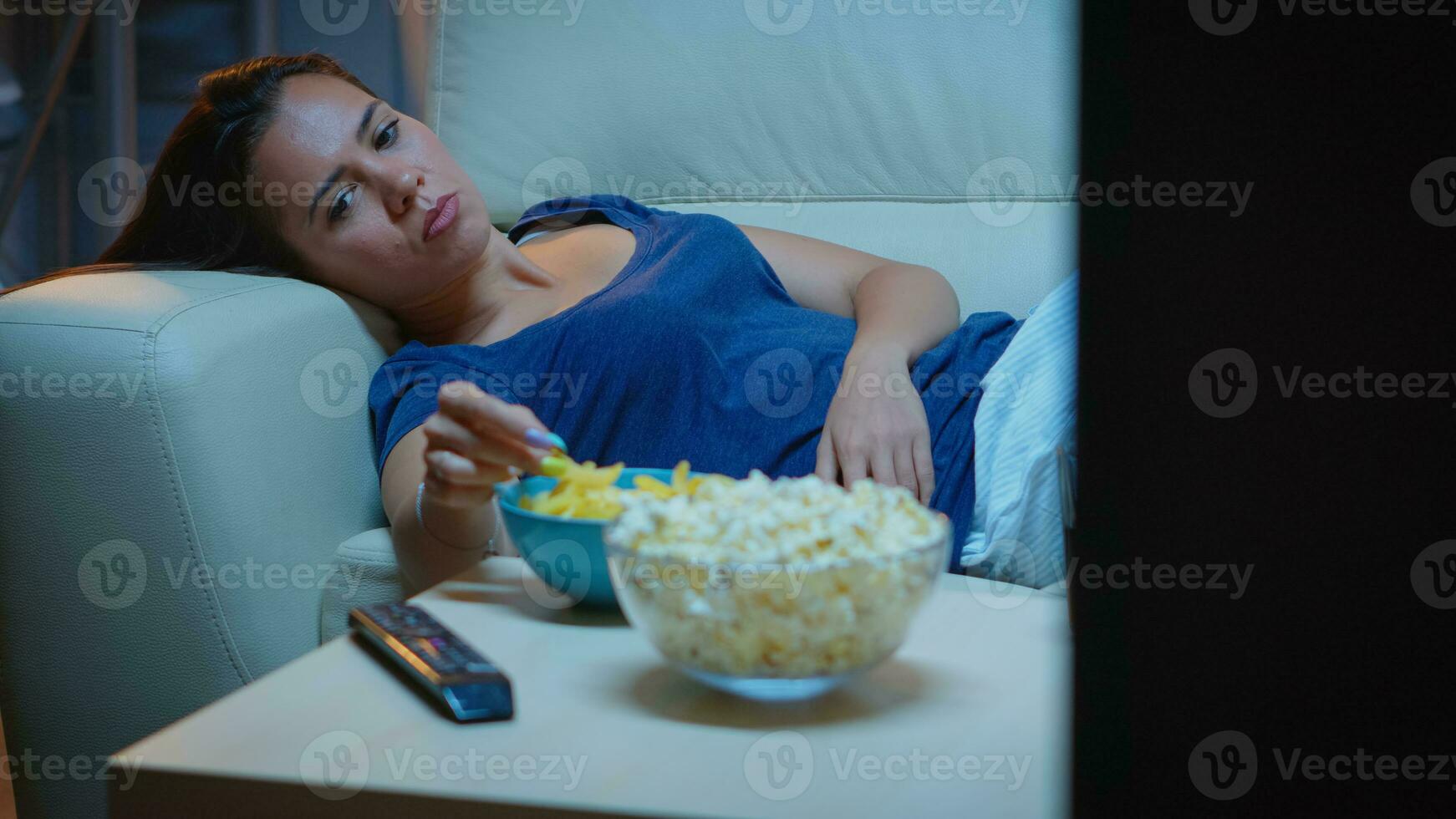 uitgeput vrouw aan het kijken TV tonen aan het liegen Aan sofa in leven kamer. moe eenzaam ongelukkig jong dame in pyjama's ontspannende Aan comfortabel bankstel in voorkant van televisie gevoel verveeld aan het eten snacks laat Bij nacht foto