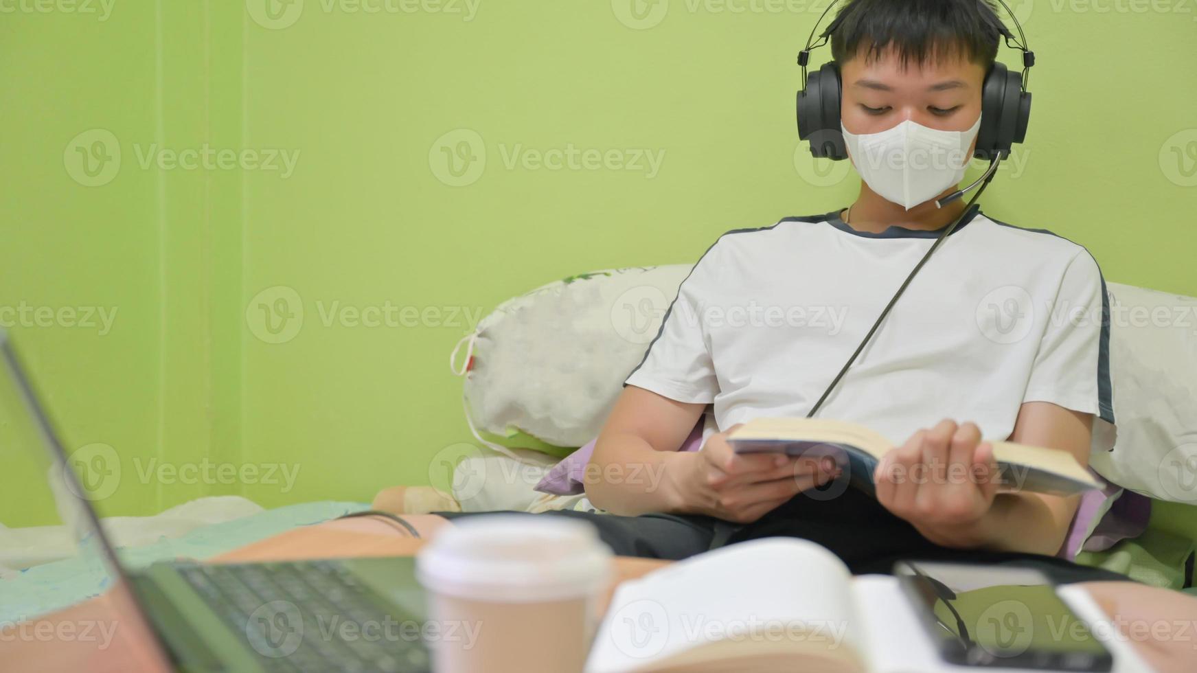 mannelijke student met masker die een boek op het bed leest om zich voor te bereiden op het examen. Hij studeerde thuis om covid-19 te beschermen. foto