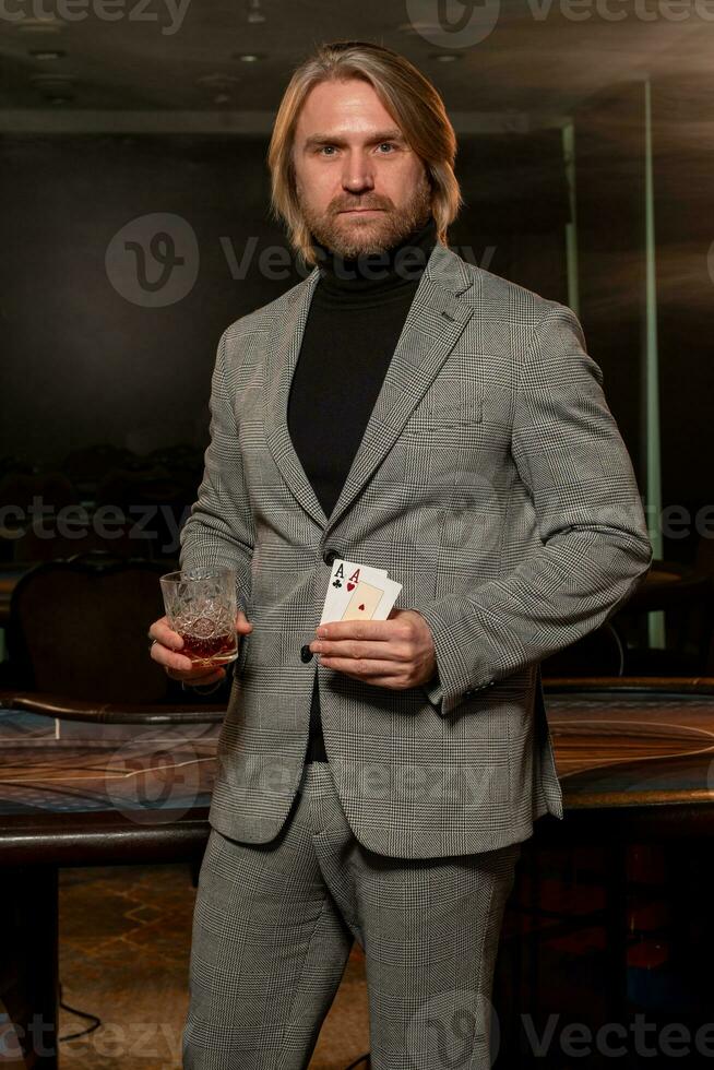 zelfverzekerd Mens staand in casino met glas van drinken en paar- van azen foto