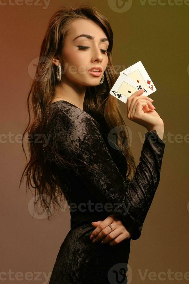brunette vrouw in zwart fluweel jurk en sieraden tonen twee azen, poseren zijwaarts Aan kleurrijk studio achtergrond. gokken, poker, casino. detailopname. foto
