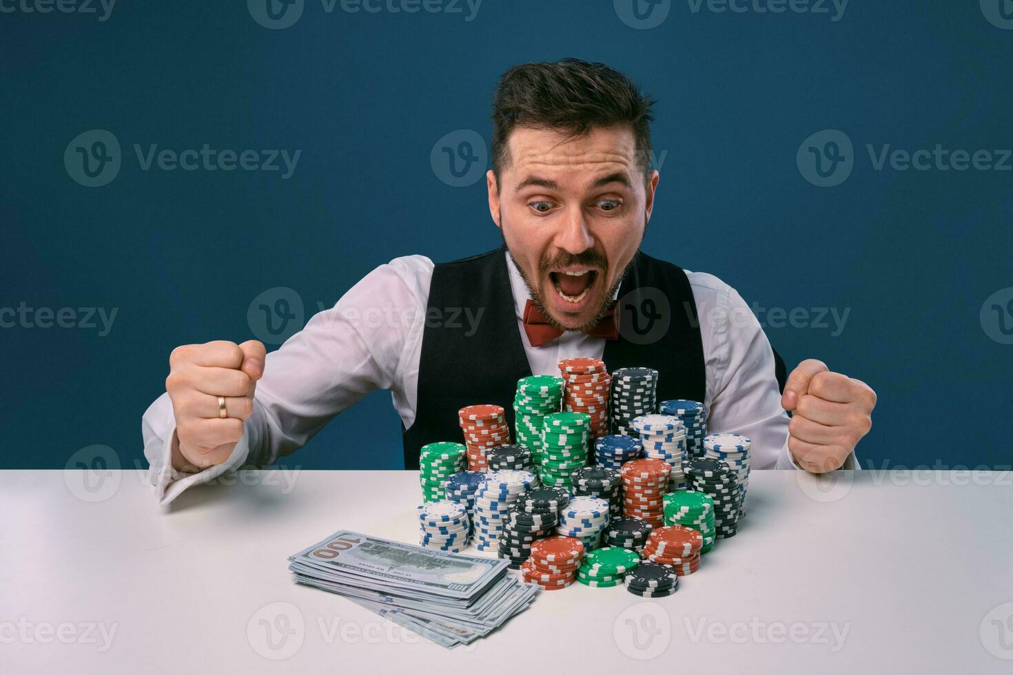 Mens in zwart hesje en overhemd zittend Bij wit tafel met stapels van chips en contant geld Aan het, poseren Aan blauw achtergrond. gokken, poker, casino. detailopname. foto