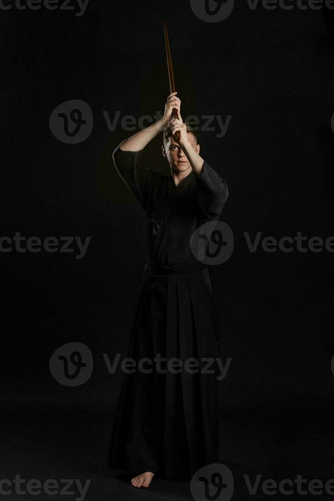 kendo goeroe vervelend in een traditioneel Japans kimono is beoefenen krijgshaftig kunst met de shinai bamboe zwaard tegen een zwart studio achtergrond. foto