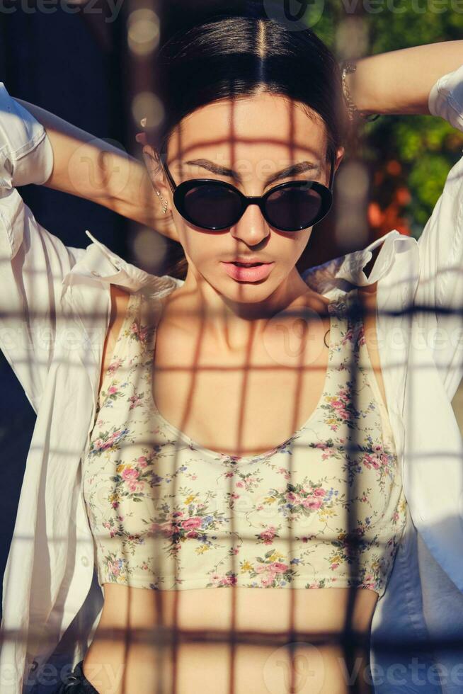 portret van meisje in zonnebril poseren in stad achter een trellized schutting. gekleed in top met bloemen afdrukken, wit shirt, zwart broek, taille tas. foto