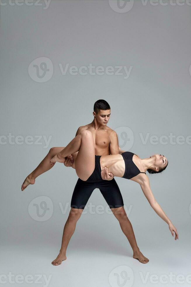 de paar van een jong modern ballet dansers in zwart pakken zijn poseren over- een grijs studio achtergrond. foto