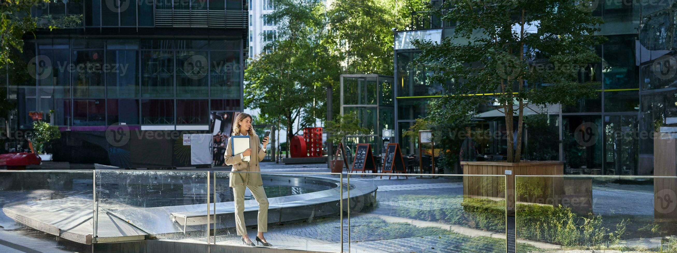 elegant zakelijke vrouw in pak, staand met laptop en werk documenten, gebruik makend van mobiel telefoon, op zoek Bij smartphone scherm foto