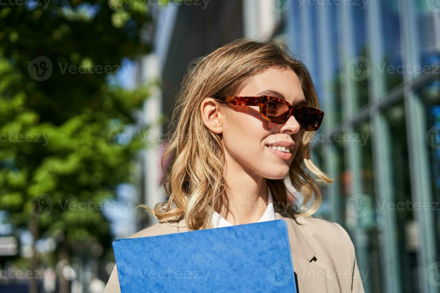 dichtbij omhoog portret van glimlachen jong vrouw in zonnebril, zakelijke pak, Holding een map met werk documenten, gaan naar een interview, staand buitenshuis foto