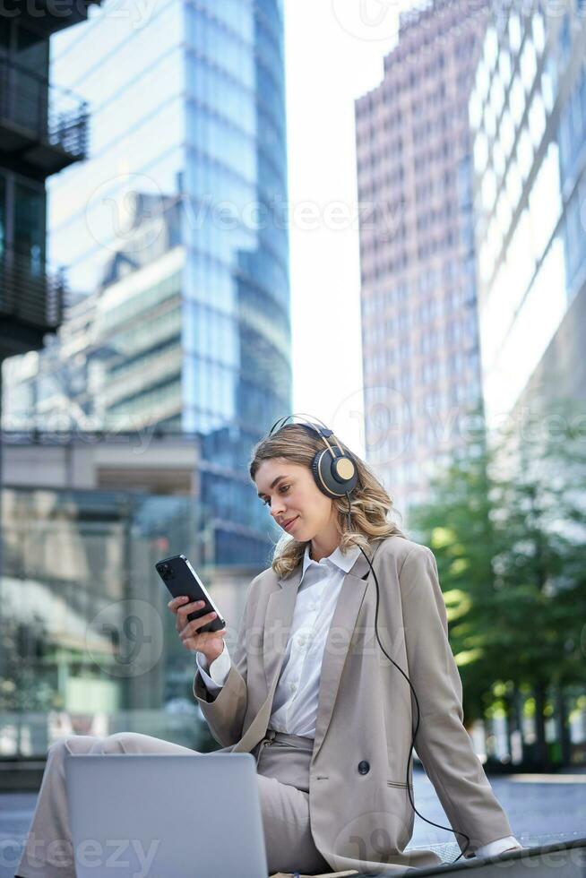 zakelijke vrouw in pak, zittend in stad, luisteren naar muziek- met laptop, scrollen nieuws Aan haar mobiel telefoon, ontspannende Aan lunch breken foto