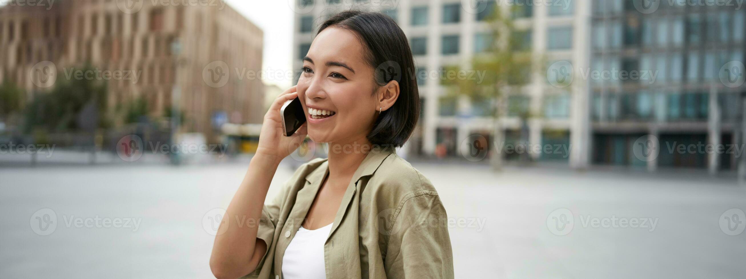 modern jong Aziatisch meisje praat Aan mobiel telefoon, toepassingen telefoon Aan stad straat. vrouw glimlachen terwijl roeping iemand Aan smartphone foto