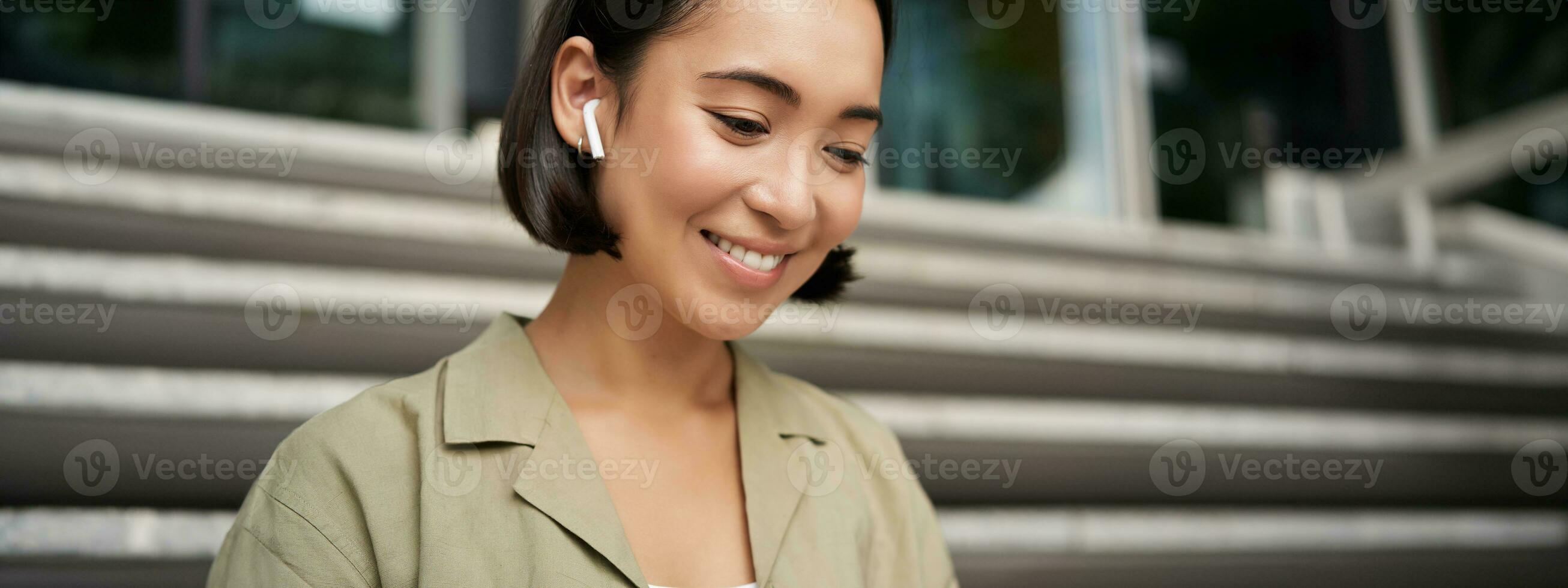 portret van glimlachen Aziatisch meisje luistert muziek, Podast in draadloze oortelefoons, gebruik makend van hoofdtelefoons buitenshuis, zittend Aan straat foto