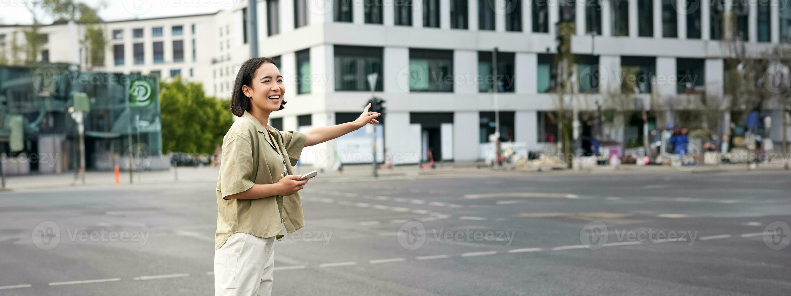 verticaal schot van jong vrouw proberen vangst taxi, golvend Bij bestuurder Aan weg, Holding smartphone met auto sharing app, staand Aan stad straat foto