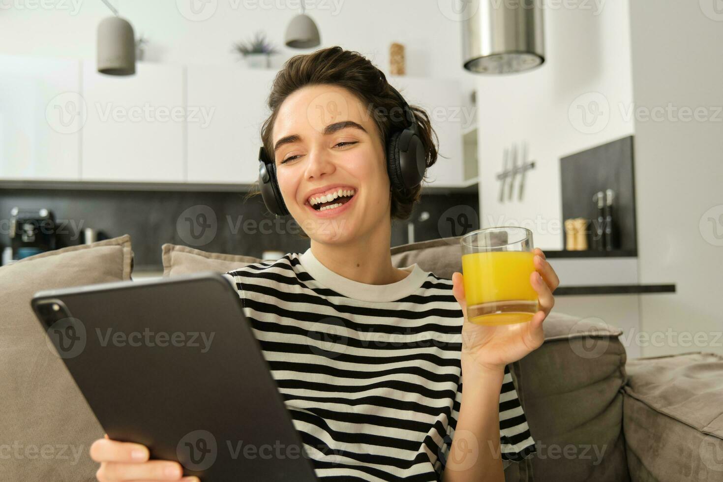 portret van vrouw in koptelefoon, zittend Aan sofa met tablet en drinken oranje sap, aan het kijken TV tonen Aan haar apparaatje, gebruik makend van sociaal media sollicitatie, uitgeven tijd Bij huis foto