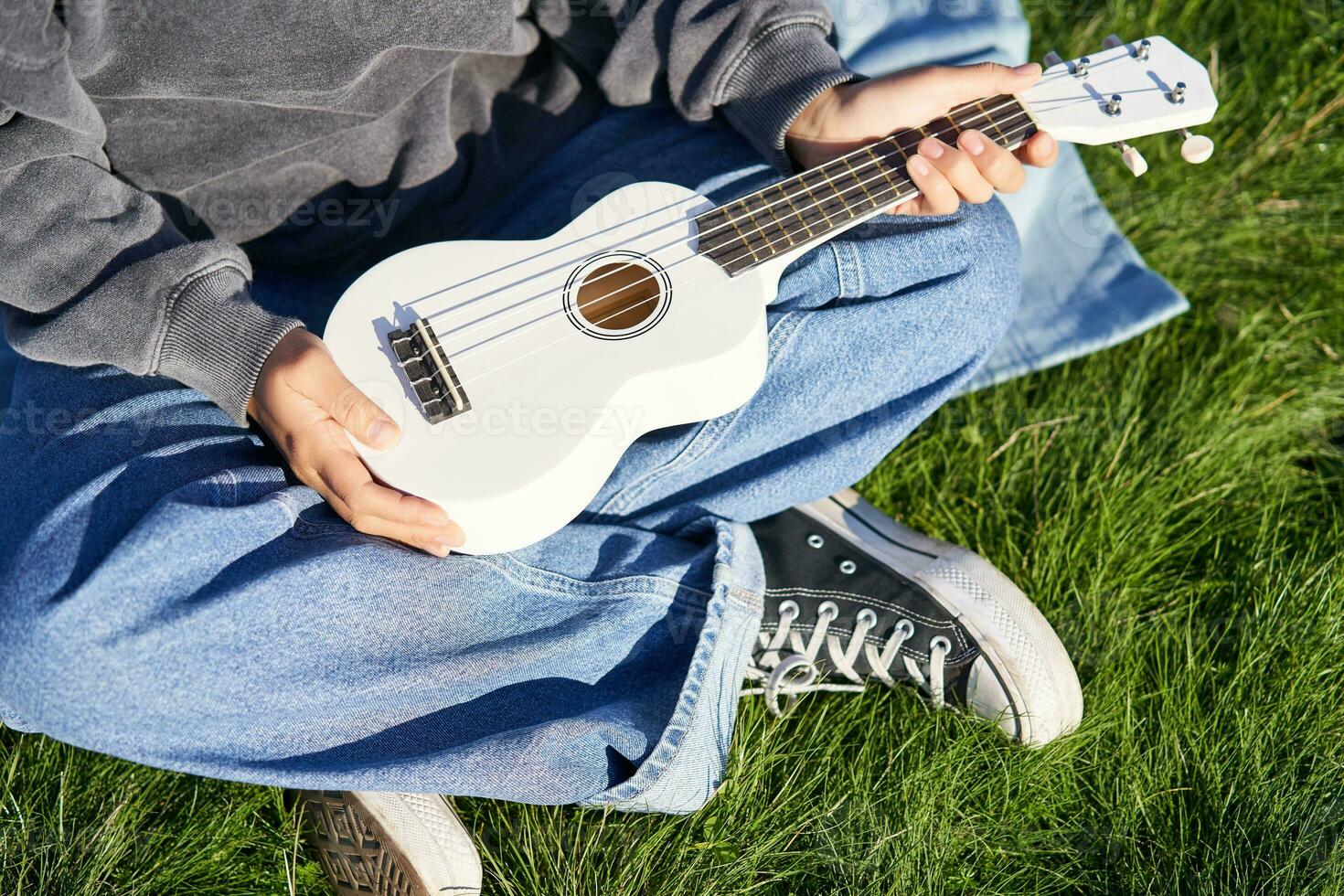 muziek- en instrumenten. dichtbij omhoog, vrouw handen Holding wit ukelele, musicus zittend Aan gras buitenshuis en spelen foto