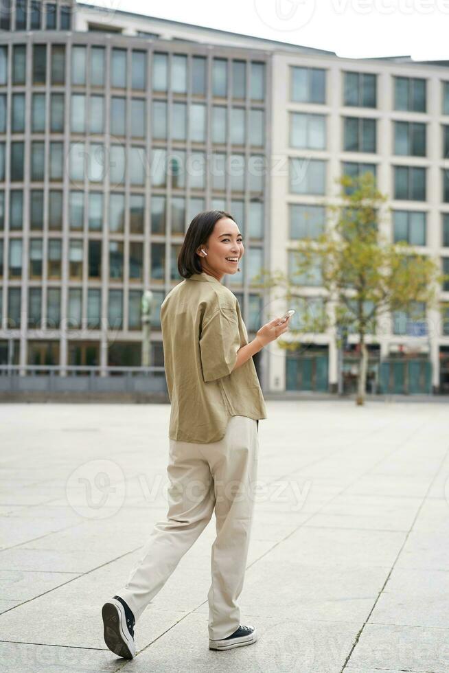 silhouet van Aziatisch vrouw wandelen Aan straat in draadloze koptelefoon, Holding smartphone foto