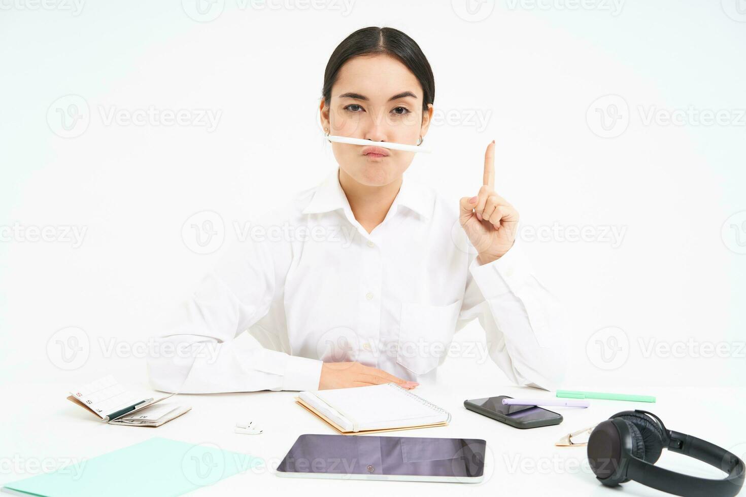 bedrijf en kantoor werk. jong vrouw zittend Bij werkplek, voor de gek houden in de omgeving van, houdt pen met lippen en shows grappig gezichten, wit achtergrond foto