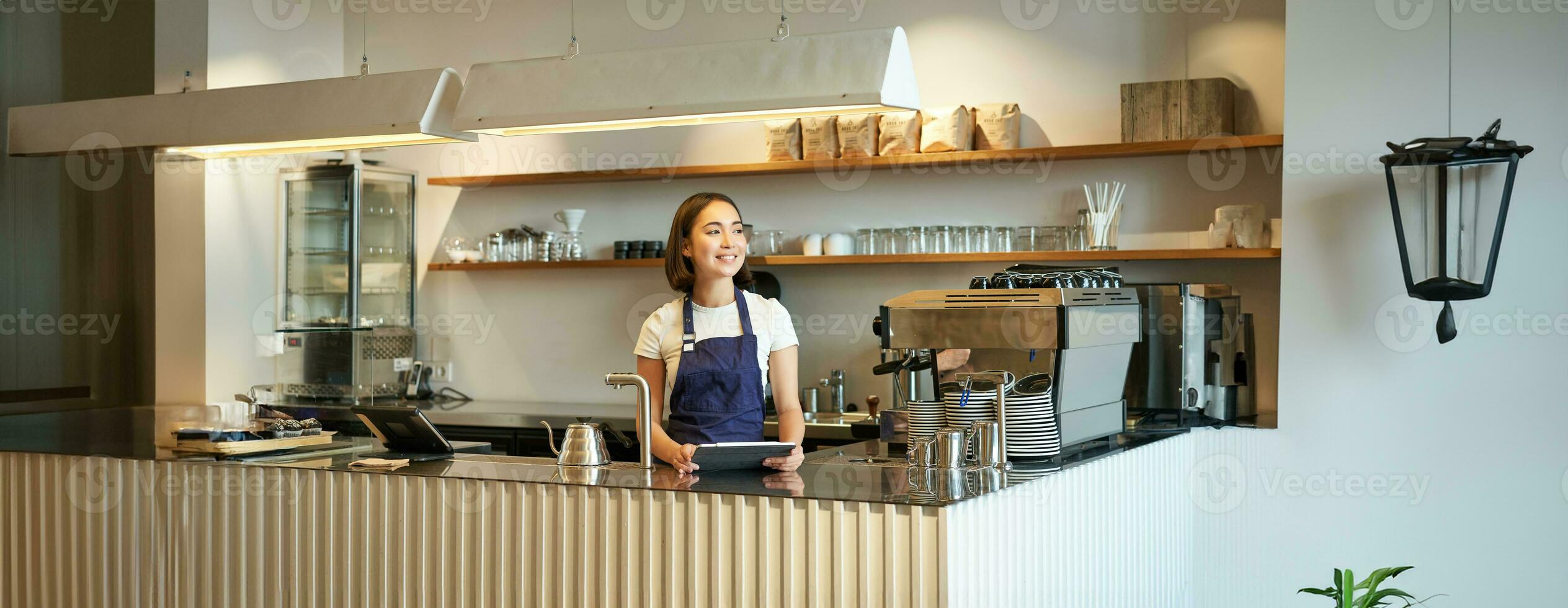 koffie winkel. glimlachen Koreaans barista, vrouw werken achter balie, gebruik makend van tablet naar nemen bestellingen in cafe, draagt blauw schort Bij werk foto