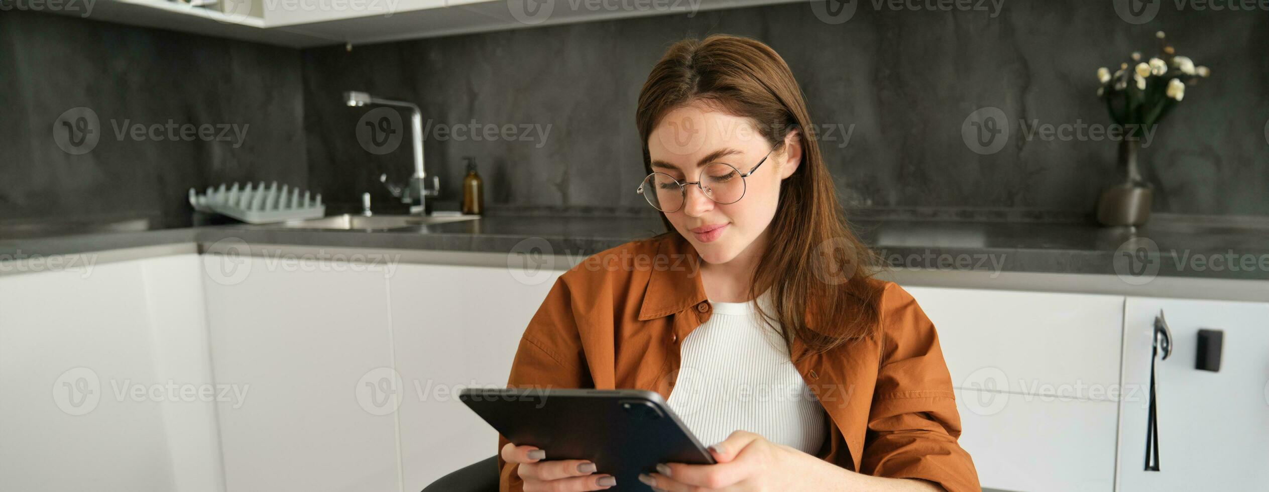 portret van brunette vrouw in bril, zittend in keuken Bij huis, Holding digitaal tablet, lezing Aan apparaatje foto