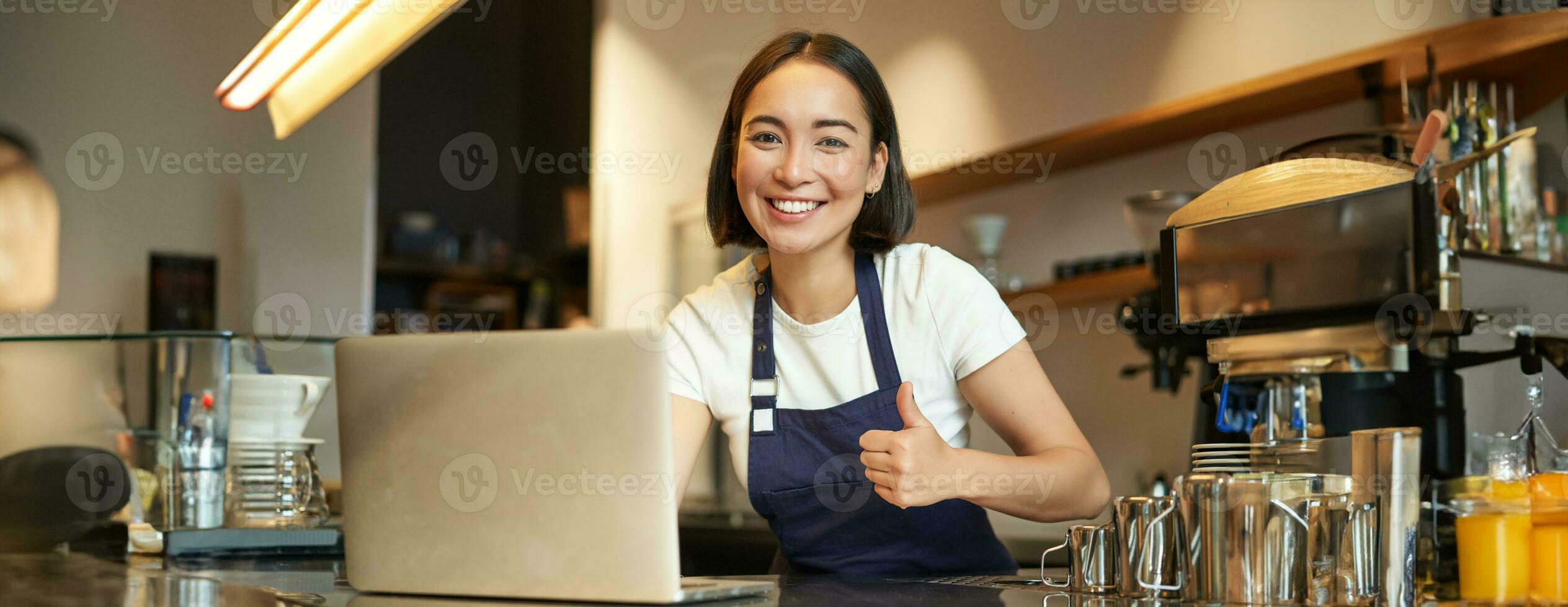 portret van Aziatisch meisje in schort, barista in koffie winkel, shows duimen omhoog, goedkeuren of adviseren iets, werken in cafe, glimlachen gelukkig foto