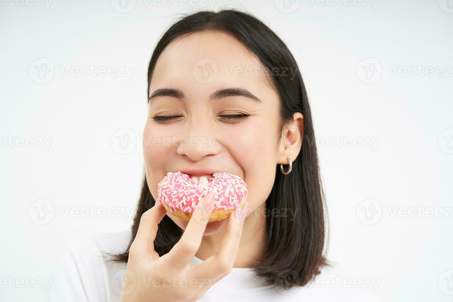 voedsel en aan het eten uit. jong Koreaans vrouw ziet eruit, duurt beet van heerlijk roze geglazuurd donut, wit achtergrond foto