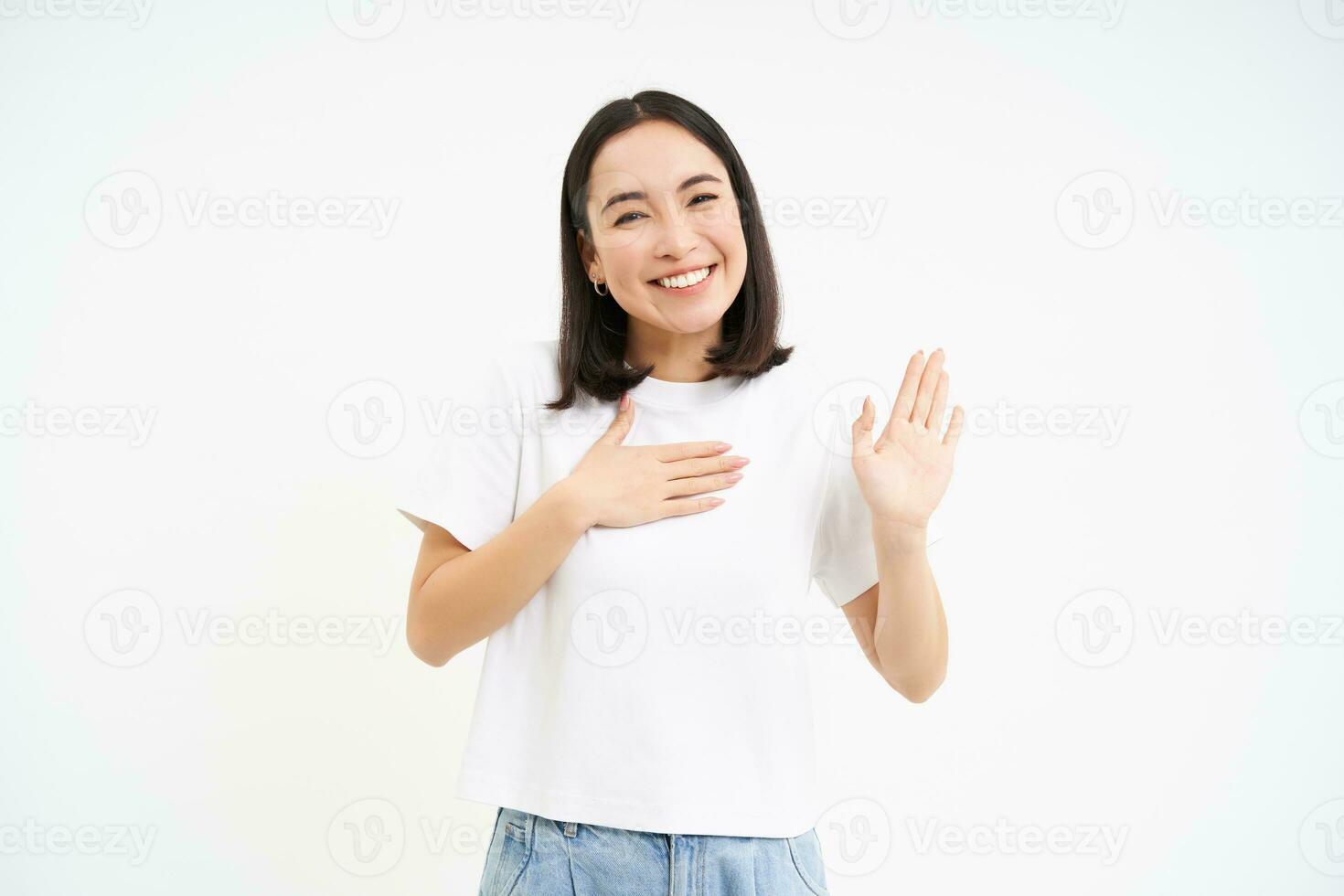 vriendelijk, glimlachen Koreaans vrouw, shows een hand- omhoog en zet palm Aan hart, maakt belofte, voorstellen haarzelf, wit achtergrond foto