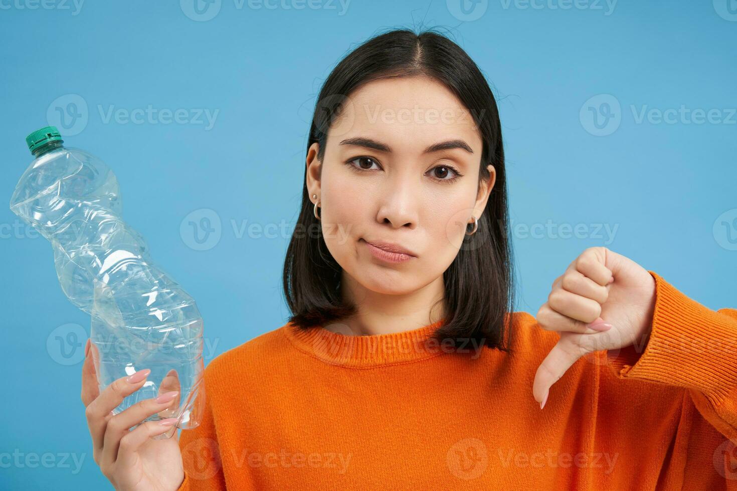 portret van Aziatisch vrouw shows duimen naar beneden en plastic fles, afkeuren gebrek van recyclen, blauw achtergrond foto
