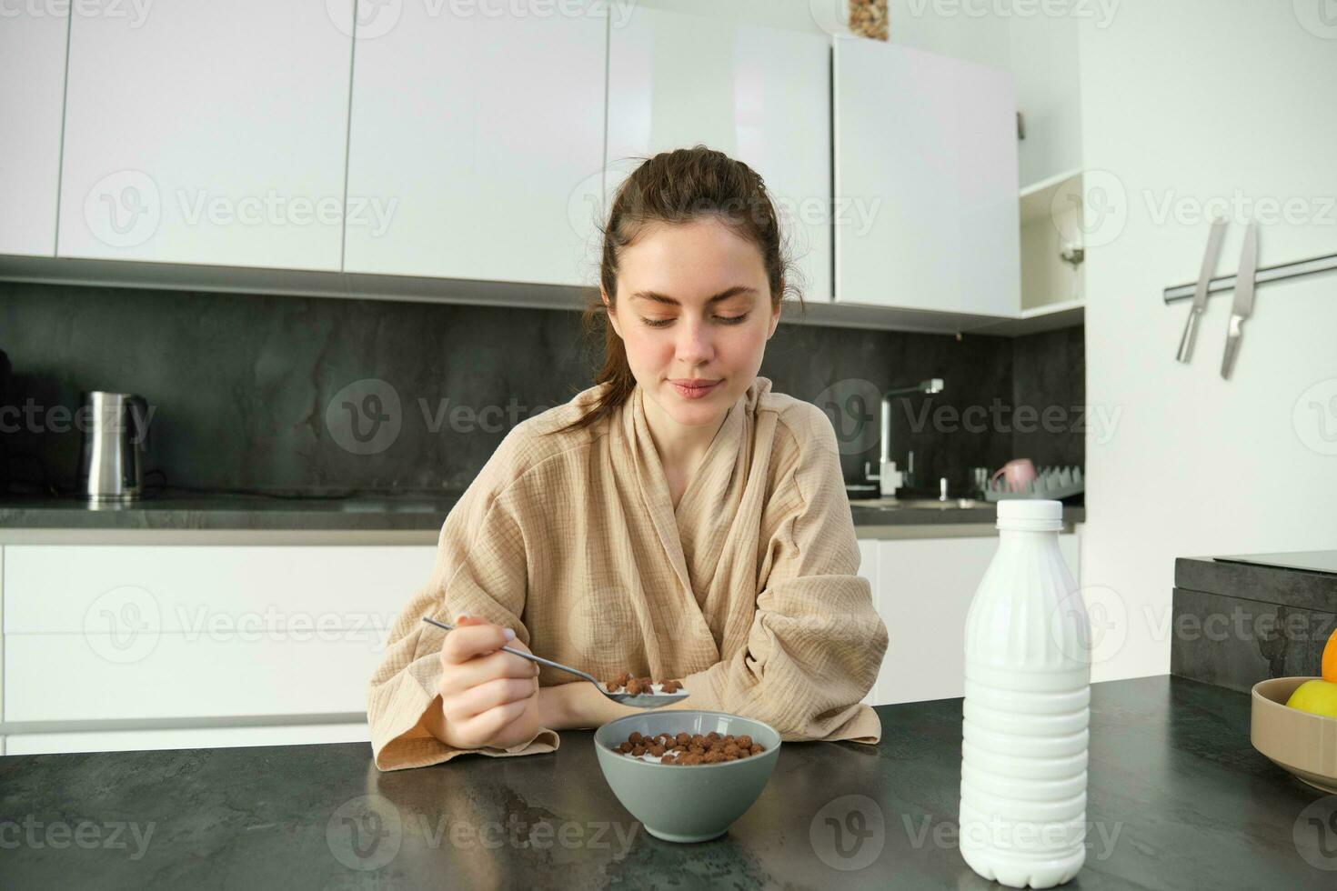 beeld van gelukkig jong vrouw aan het eten granen Bij huis met melk, hebben haar ontbijt, vervelend badjas, zittend in keuken alleen foto