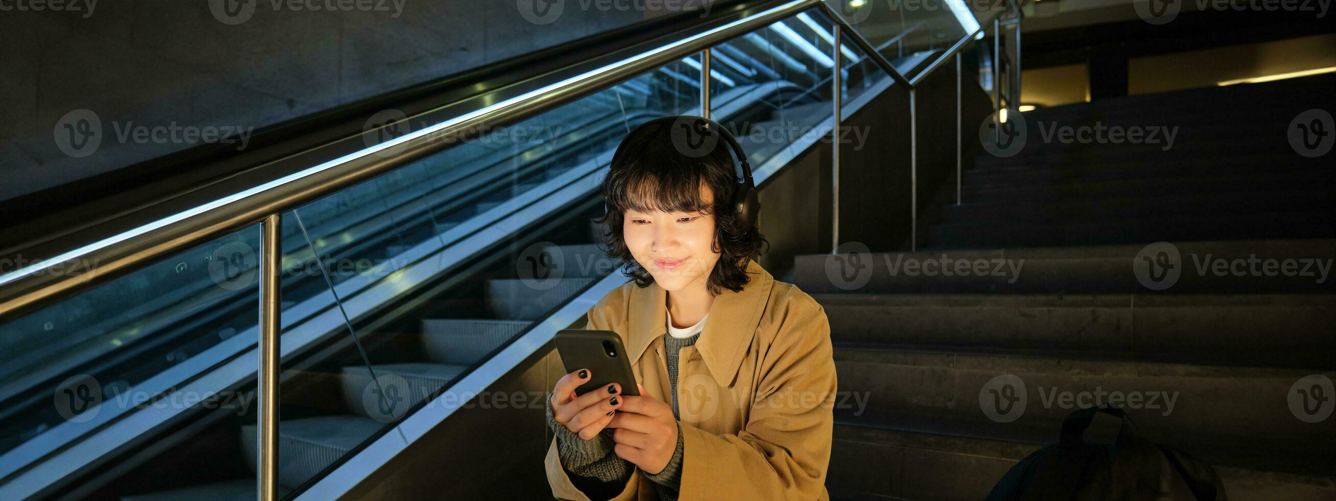technologie. portret van Aziatisch meisje zit Aan trap in de buurt roltrap, luistert muziek- in hoofdtelefoons en toepassingen sociaal media, leest, horloges video in openbaar plaats foto