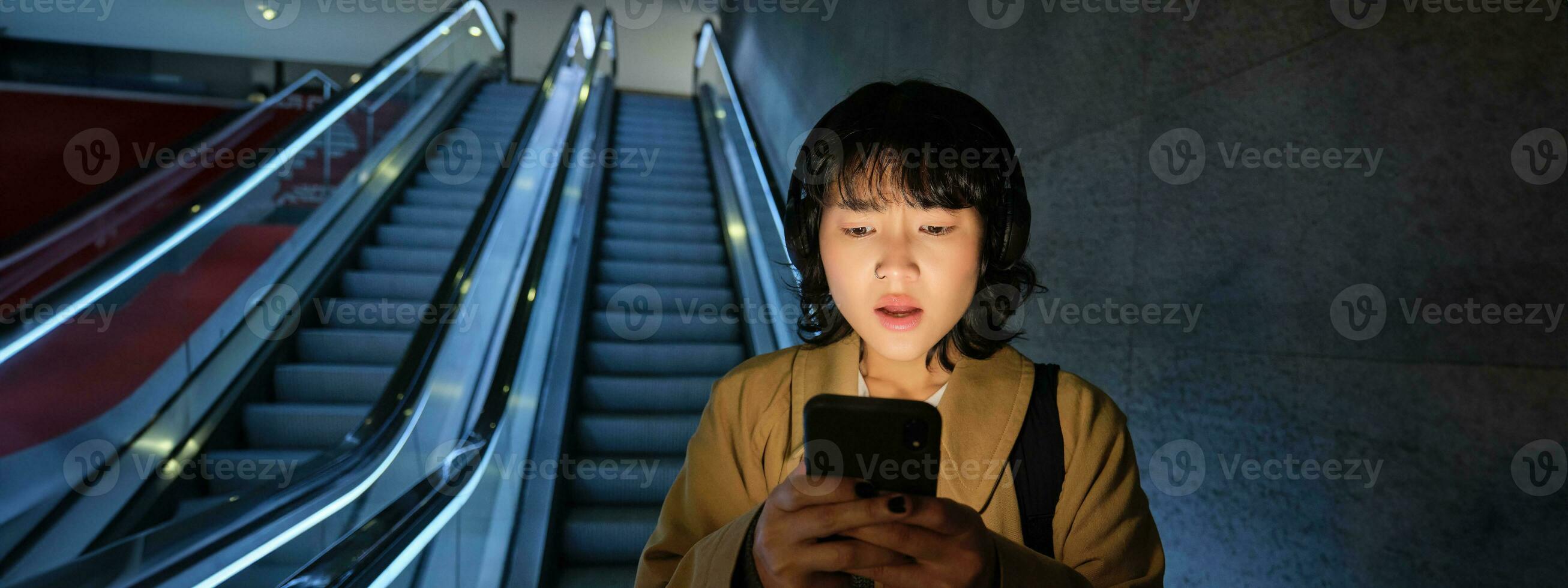 portret van Aziatisch meisje looks geschokt Bij mobiel telefoon scherm, staand Aan roltrap, gaan naar beneden naar ondergronds of metro, woon-werkverkeer naar werk foto