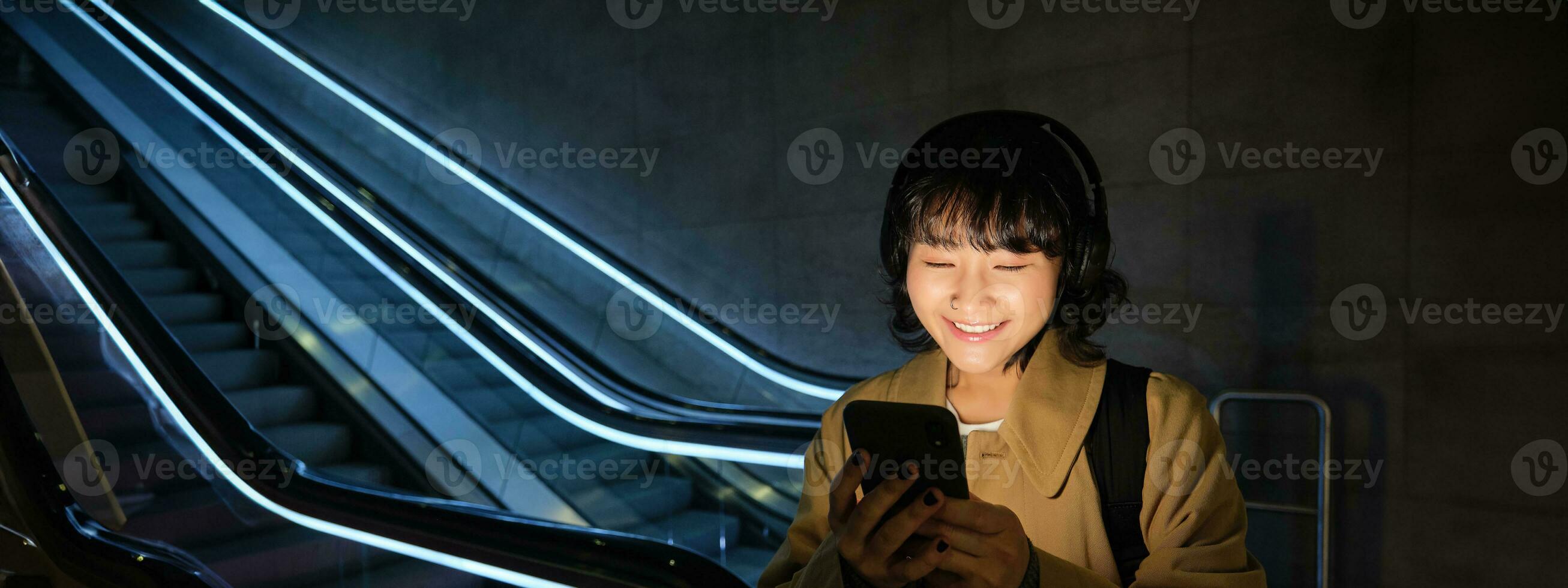 gelukkig Aziatisch vrouw in metro, staand in de buurt roltrap, Holding smartphone en luisteren muziek- in koptelefoon, glimlachen verheugd foto