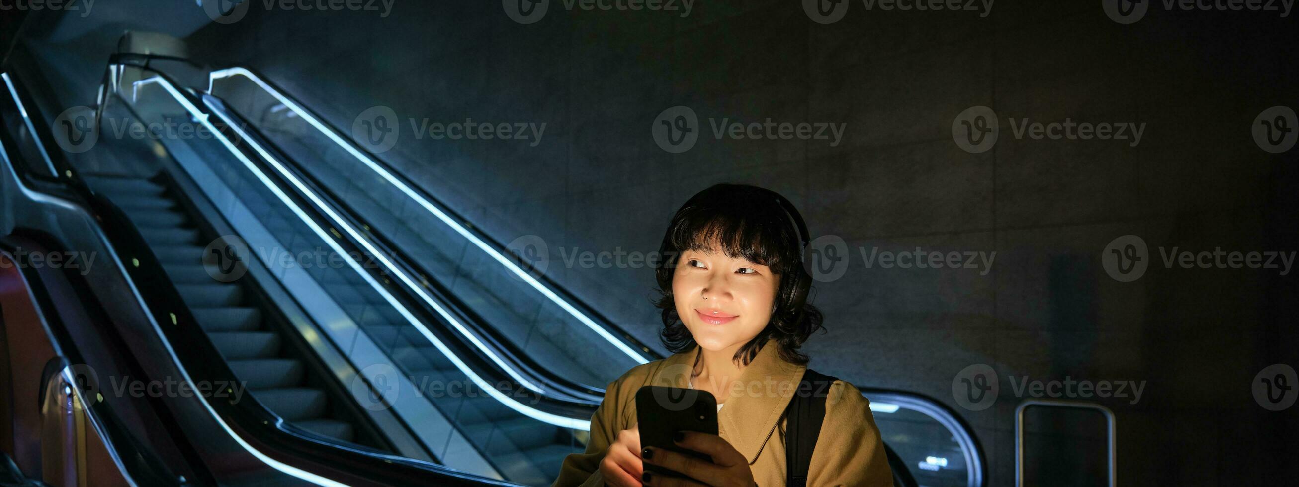 glimlachen Koreaans meisje in koptelefoon, luistert muziek, toepassingen mobiel telefoon in buis, staat in de buurt roltrap in donker, looks vrolijk foto