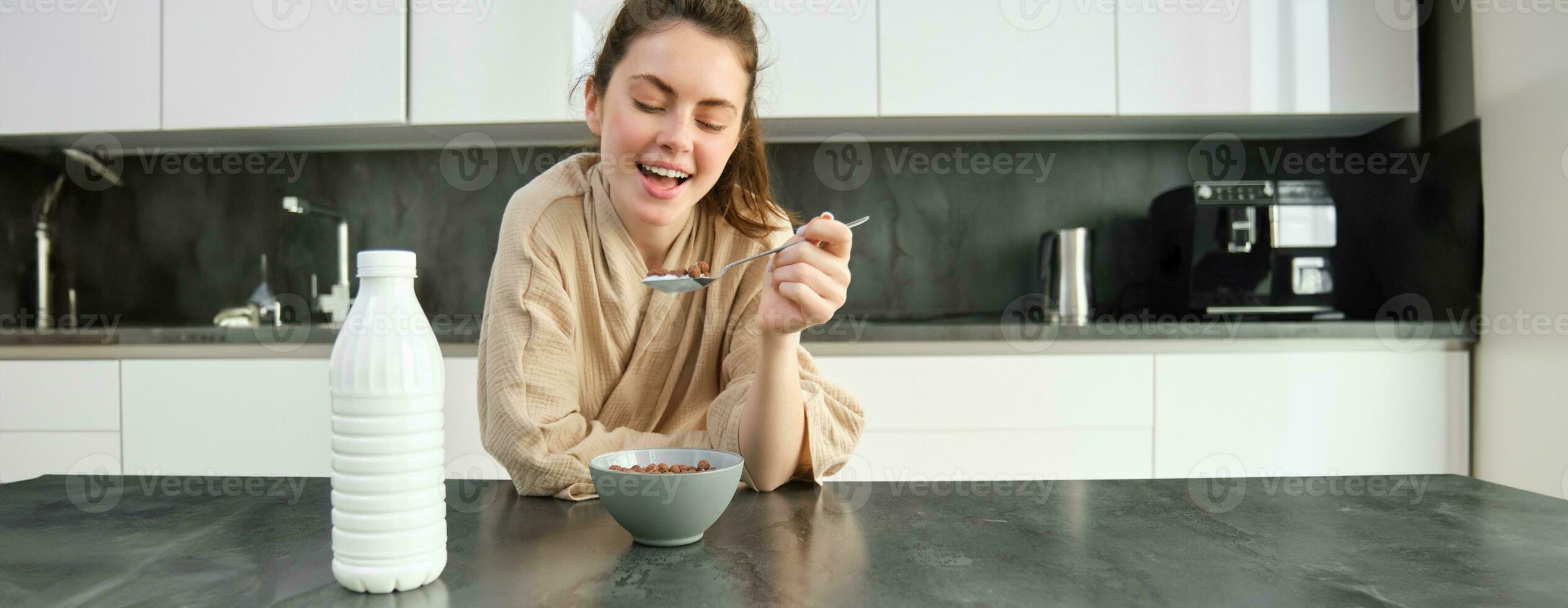 portret van mooi jong en gezond vrouw in badjas eet haar ontbijt in keuken, heeft granen met melk en glimlachen foto