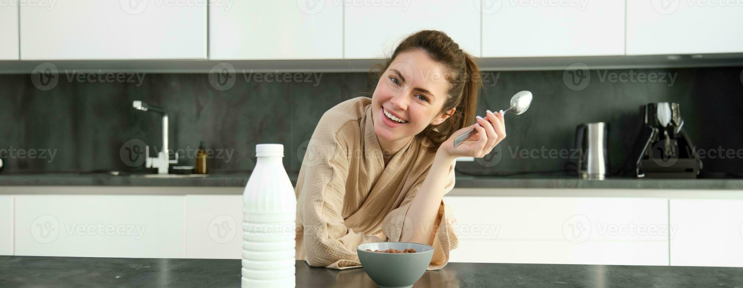 portret van jong mooi vrouw in badjas, aan het eten granen voor ontbijt, leunt Aan keuken werkblad, op zoek Bij haar ochtend- maaltijd foto