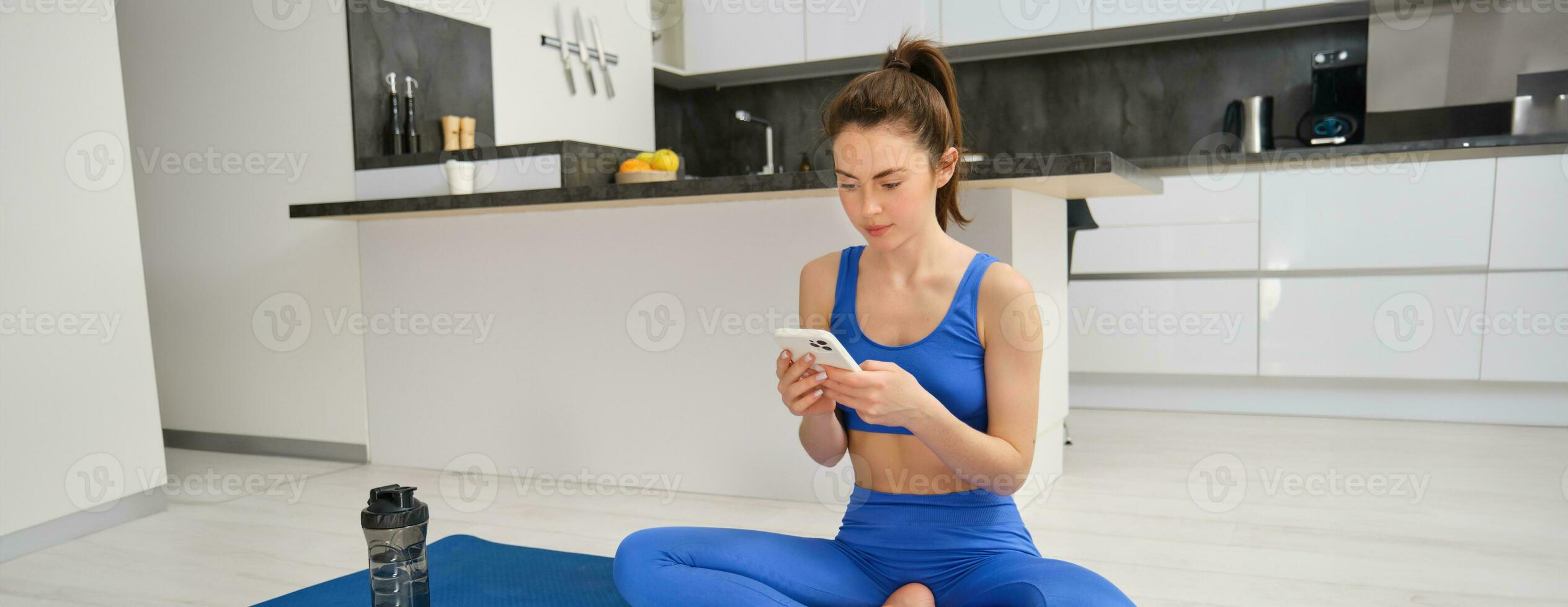 portret van vrouw gebruik makend van geschiktheid app, aan het doen training van huis, zit Aan yoga mat met smartphone in leven kamer foto