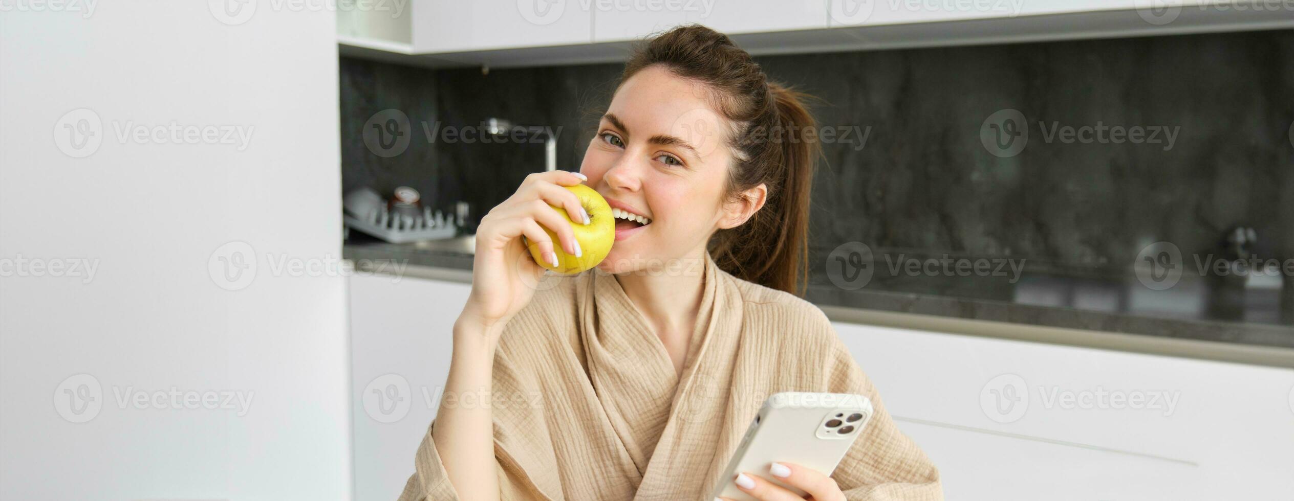 dichtbij omhoog portret van glimlachen mooi vrouw, zittend in badjas, aan het eten appel en op zoek Vrolijk, gebruik makend van smartphone terwijl zittend in de keuken, bestellen voedsel online Aan mobiel app foto