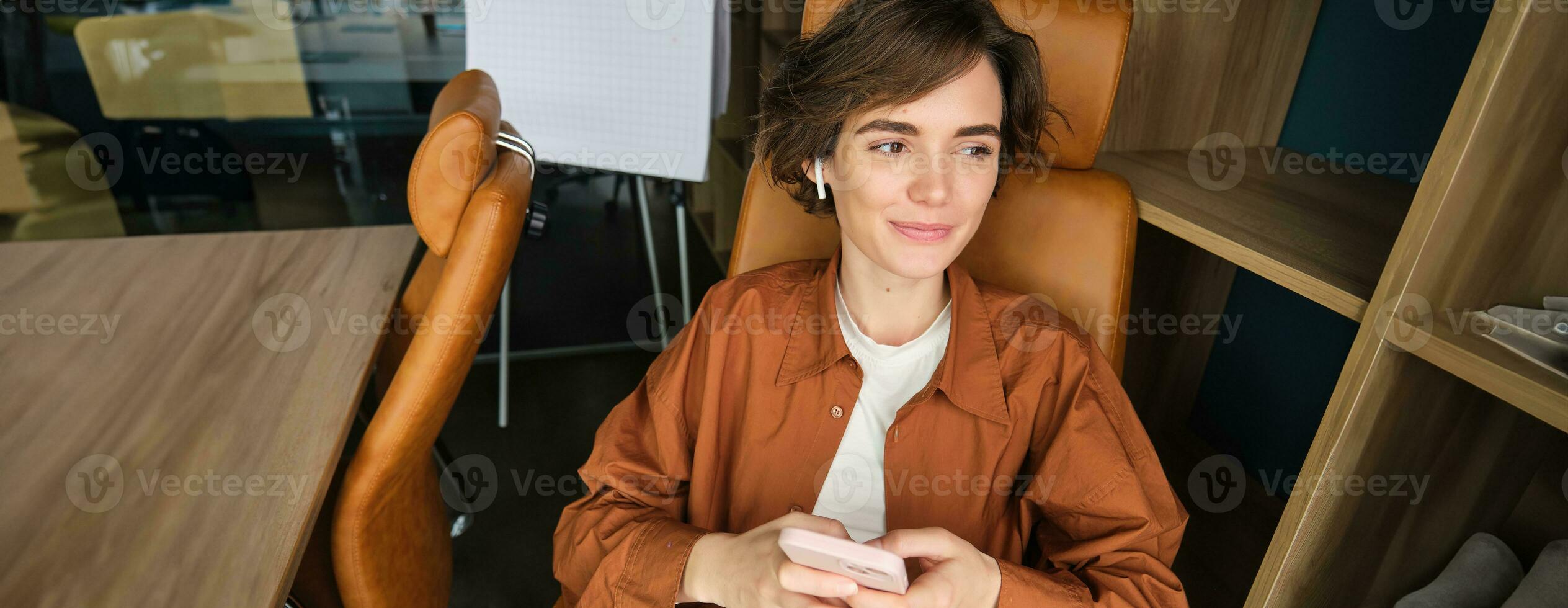 portret van vrouw werken in een kantoor, werknemer gebruik makend van smartphone, zittend in stoel Aan haar breken en glimlachen foto