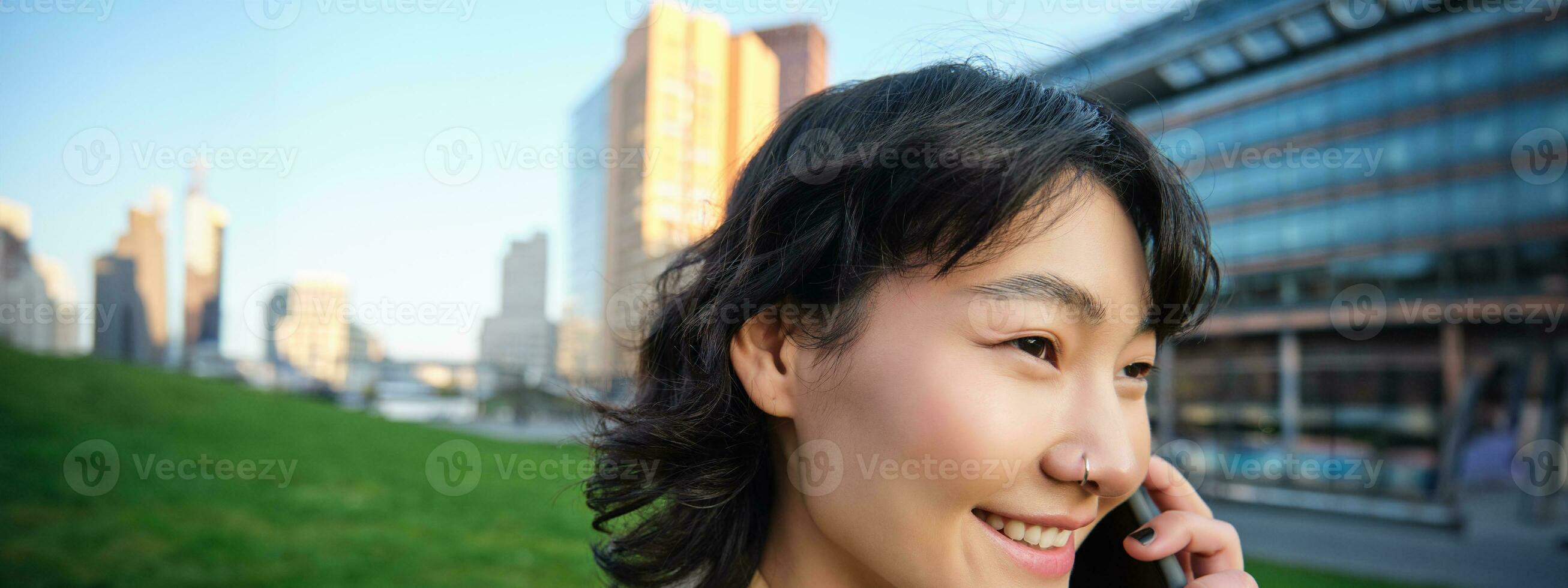 hoofdschot van jong Koreaans vrouw wandelingen Aan straat, praat Aan mobiel telefoon, heeft telefoon gesprek, staat in de buurt campus gebouw foto