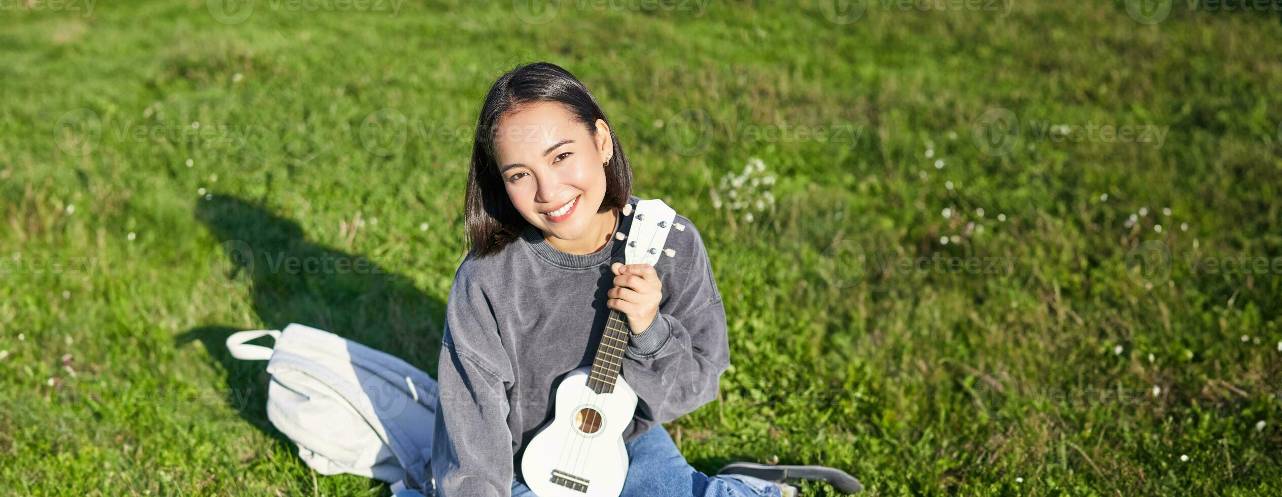 glimlachen Aziatisch meisje leert hoe naar Speel ukulele Aan laptop, video babbelen met muziek- docent, zittend met instrument in park Aan gras foto