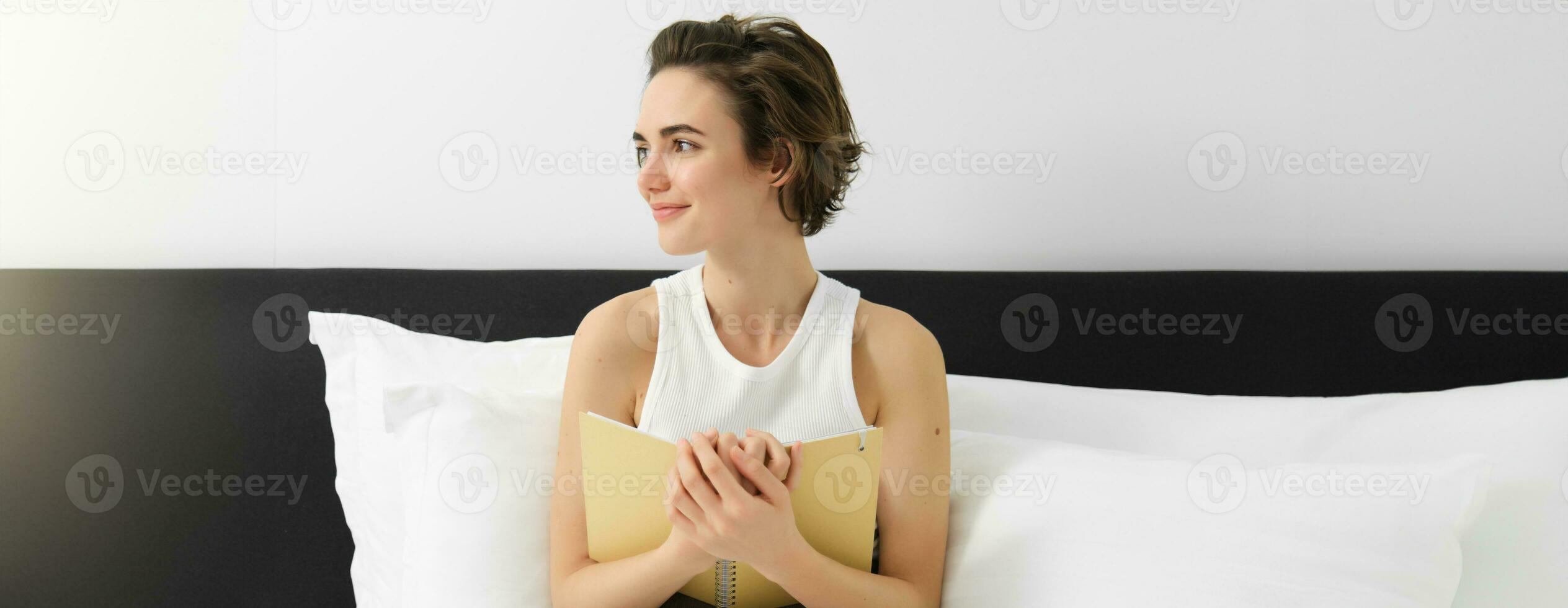 portret van jong sensueel vrouw, knuffelen haar notitieboekje, schrijven in logboek of dagboek, glimlachen en op zoek terzijde, zittend Aan bed, opmerkend haar dromen of gevoelens in ochtend- foto